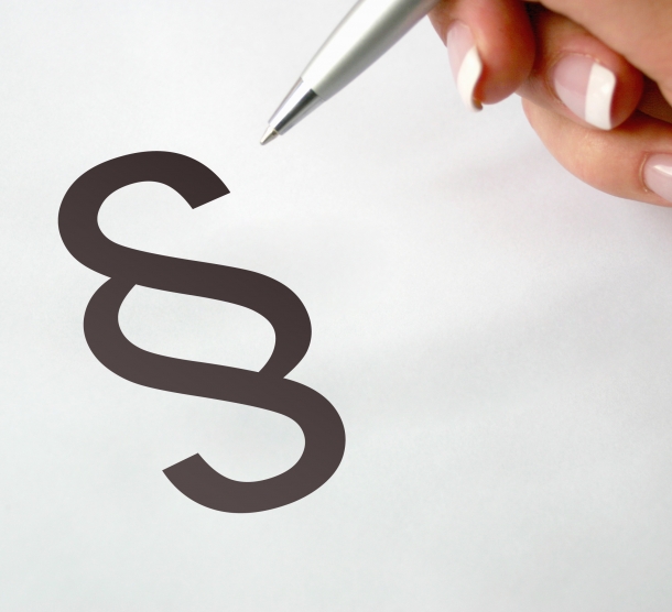 Ein großes Paragraphen-Symbol auf weißem Grund. Die Hand einer Frau hält einen Kugelschreiber, der auf das Symbol zeigt.