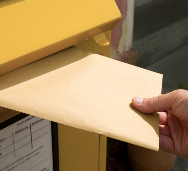 Eine Hand, die ein scheinbar unbeschriftetes Briefkuvert in einen öffentlichen Brifkasten einsteckt.